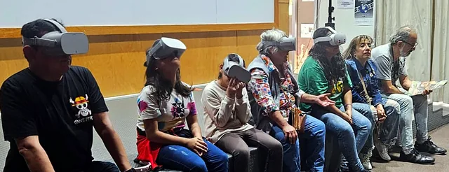 Niñas, niños y adultos sentados en un escenario, con visores de realidad virtual